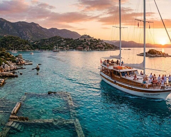 A boat named 'NEOLIA GOVERIGRE' on a clear turquoise sea with rocky outcrops and a mountainous backdrop during sunset. 4