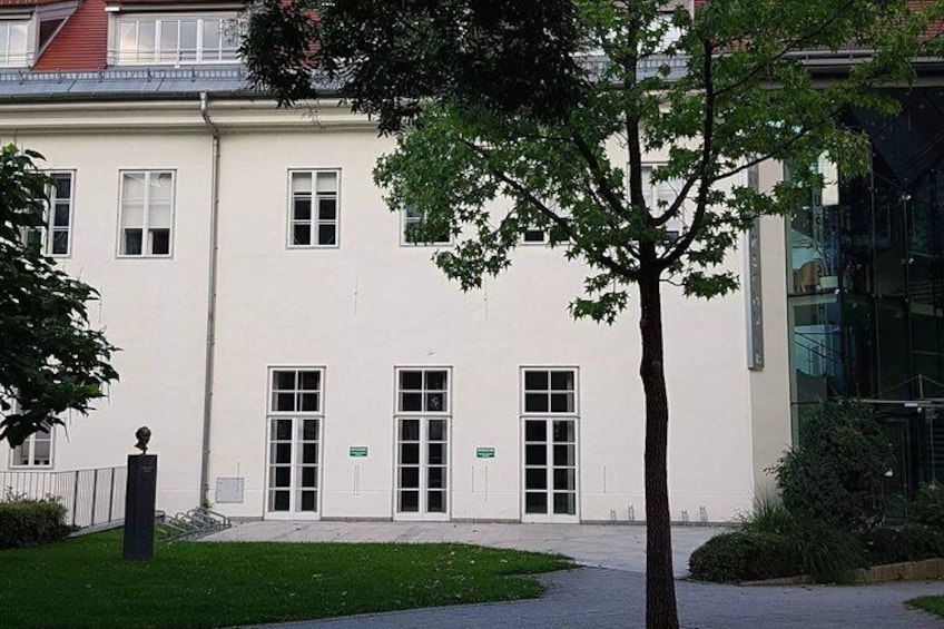 A white building with multiple windows and doors, surrounded by a small lawn and a tree. 1
