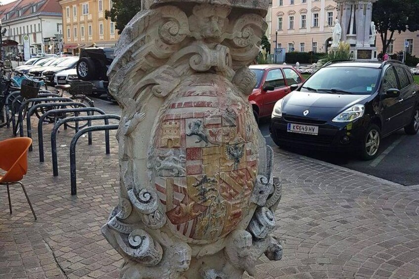 A stone monument with intricate carvings and a mosaic, surrounded by parked cars and a chair on a cobblestone street. 5