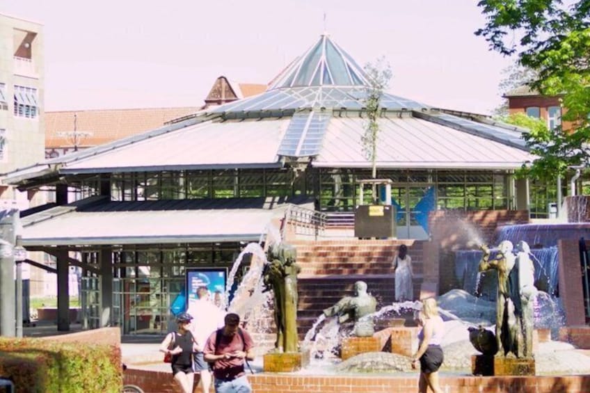 A building with a glass facade, a fountain with statues, and a tree in front. 1