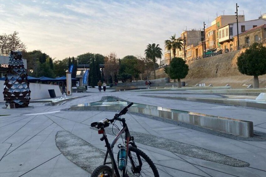 A bicycle parked in a public square with a fountain and buildings in the background. 4