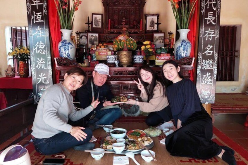 A group of people sitting around a table with food and drinks, in a room with traditional decorations and a shrine in the background. 3