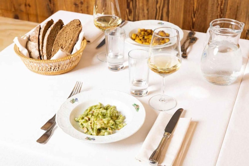 A table set with a plate of green pasta, a basket of bread, wine glasses, and a pitcher of water. 5