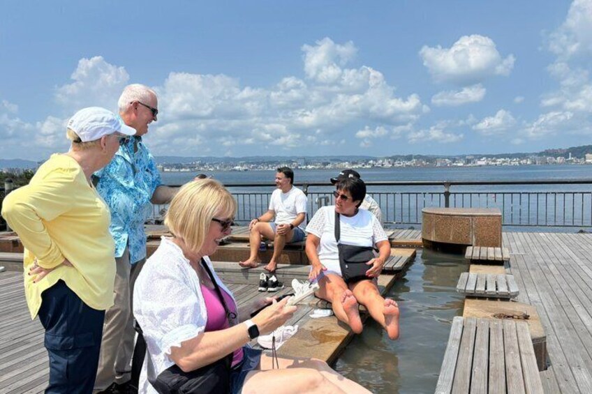 A group of people on a wooden dock by a body of water, some sitting and others standing, with a cityscape in the background. 5
