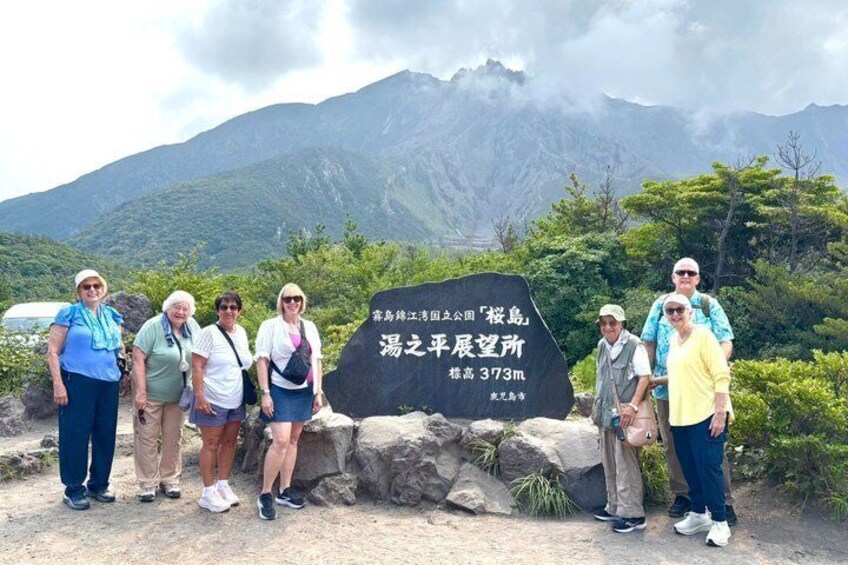 A group of people standing in front of a sign with text in a foreign language, with a mountainous landscape in the background. 1
