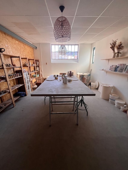 A workshop with a large work table, shelves with pottery, and a hanging light fixture. 1