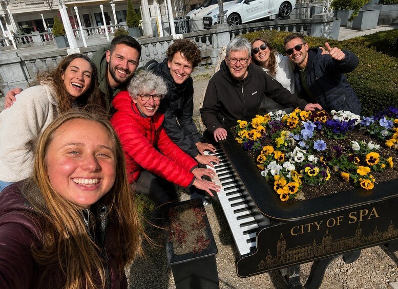 A group of people gathered around a piano with flowers on it, with a building and parked cars in the background. 1