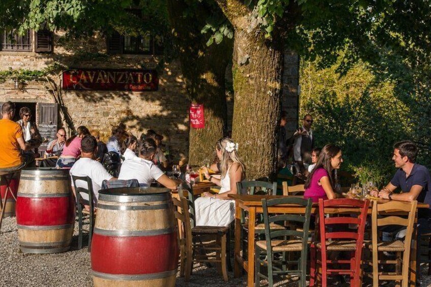 Outdoor seating at a restaurant with patrons dining, barrels, and a building with a sign. 2
