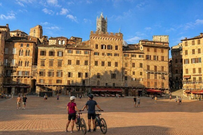 A historic square with buildings, a prominent tower, and people walking and cycling. 3