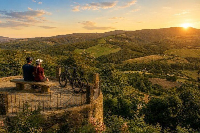A couple sitting on a bench overlooking a hilly landscape with a bicycle leaning against a fence. 1