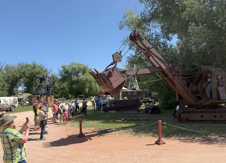 A large, rusted mechanical structure with a wheel and a long arm, surrounded by people and vehicles in a park-like setting. 4