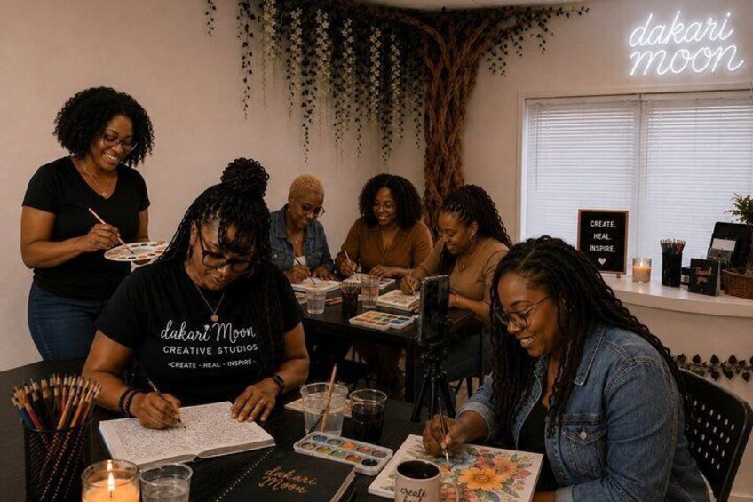 A group of people engaged in a creative activity at dakari moon creative studios, with art supplies and a sign that reads 'CREATE. HEAL. INSPIRE.' 3