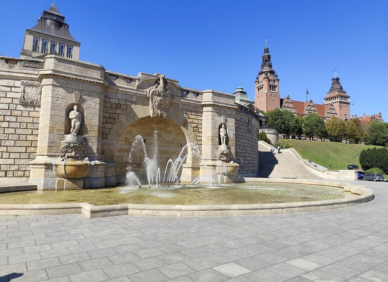 A historic fountain with statues, a stone archway, and a building with a tower in the background. 4