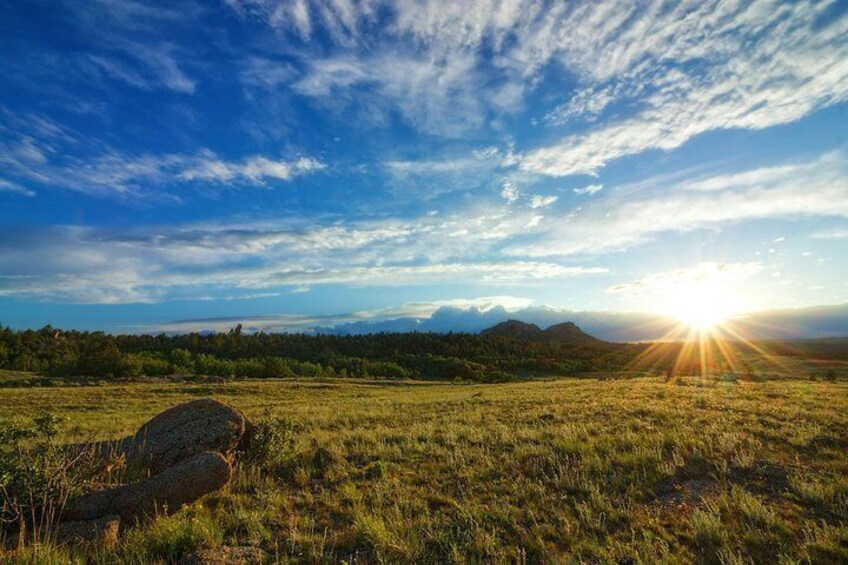 A grassy field with a large rock and mountains in the background under a blue sky with the sun setting. 4