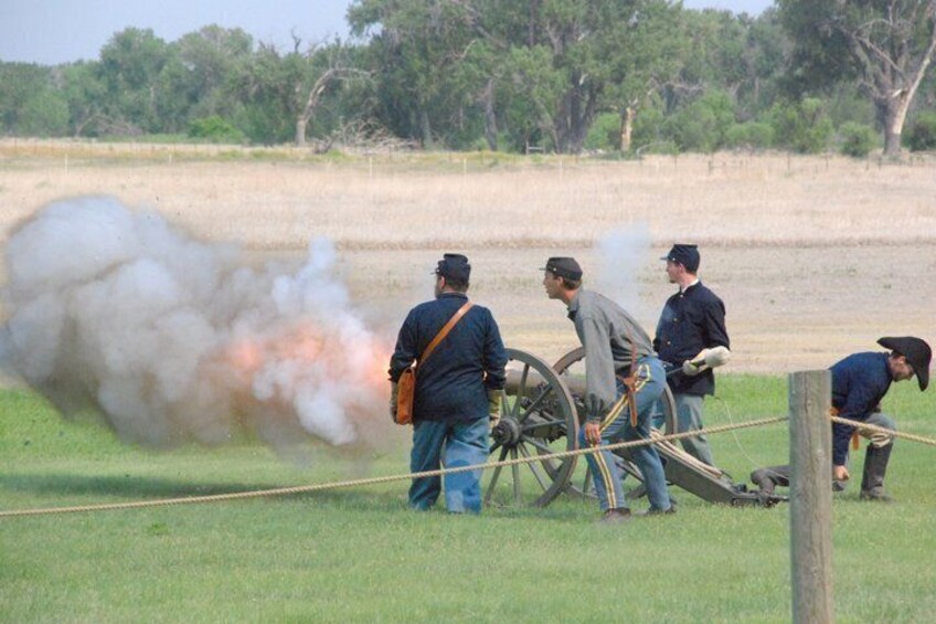 A historical reenactment with a cannon firing, surrounded by participants in period uniforms. 1