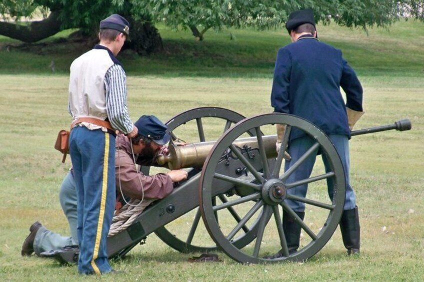 A historical reenactment with a man in a cannon carriage, another man aiming a cannon, and a third man standing by. 2