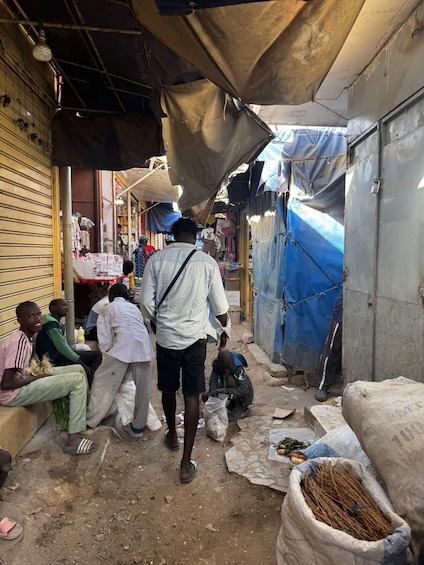 A bustling market street with people walking and sitting, covered stalls, and various goods displayed. 5