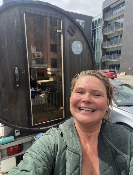 A person in a green jacket standing next to a dark-colored trailer with a glass door, parked in front of a building with multiple windows and balconies. 2