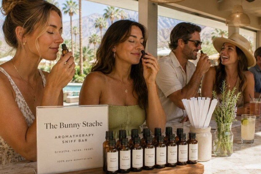 A group of people at a table with a sign reading 'The Bunny Stache Aromatherapy Sniff Bar' and bottles of essential oils. 1