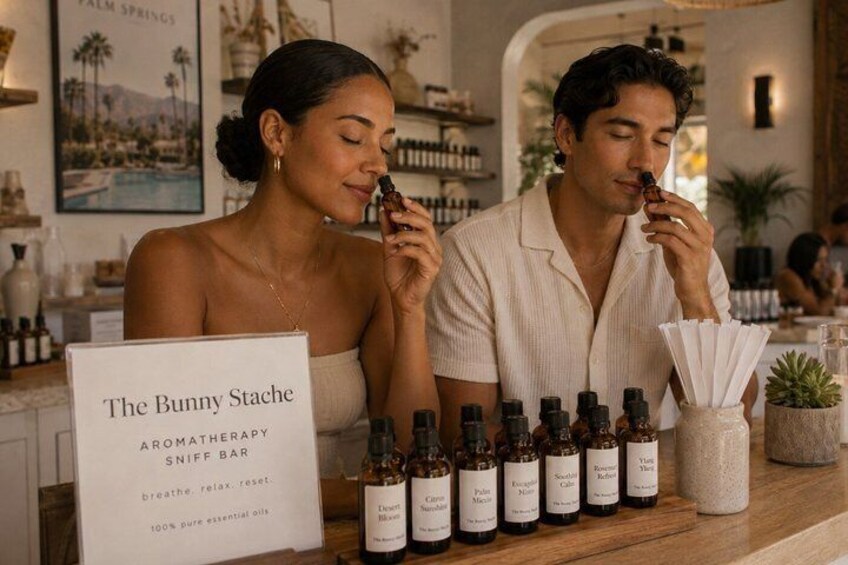 A couple at a bar with a sign reading 'The Bunny Stache Aromatherapy Sniff Bar' and various bottles of essential oils. 2