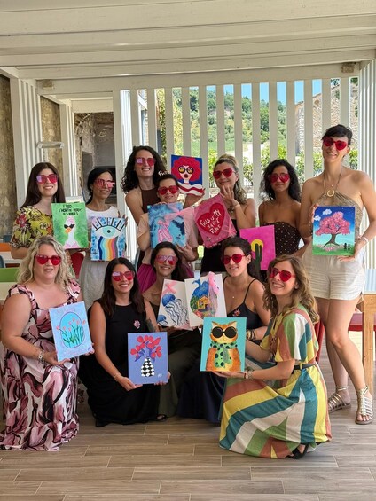 A group of people holding up colorful paintings in a covered outdoor area with a wooden fence and greenery in the background. 3