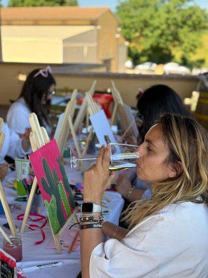 A person holding a glass, with art supplies and paintings on a table. 2