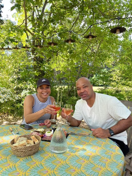 Two people toasting with drinks at an outdoor table surrounded by greenery. 3