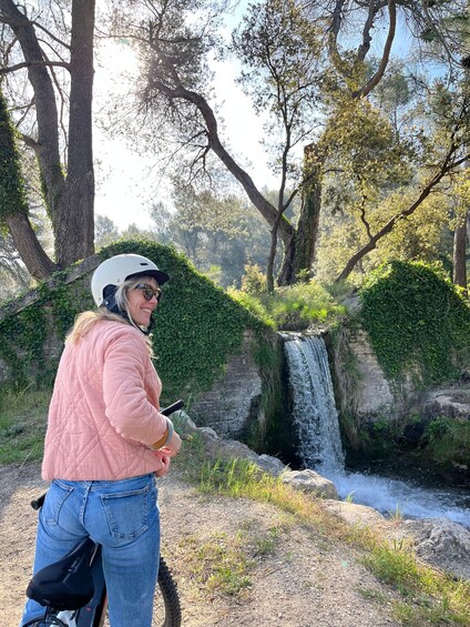 A person in a pink jacket and helmet near a waterfall surrounded by trees and greenery. 1