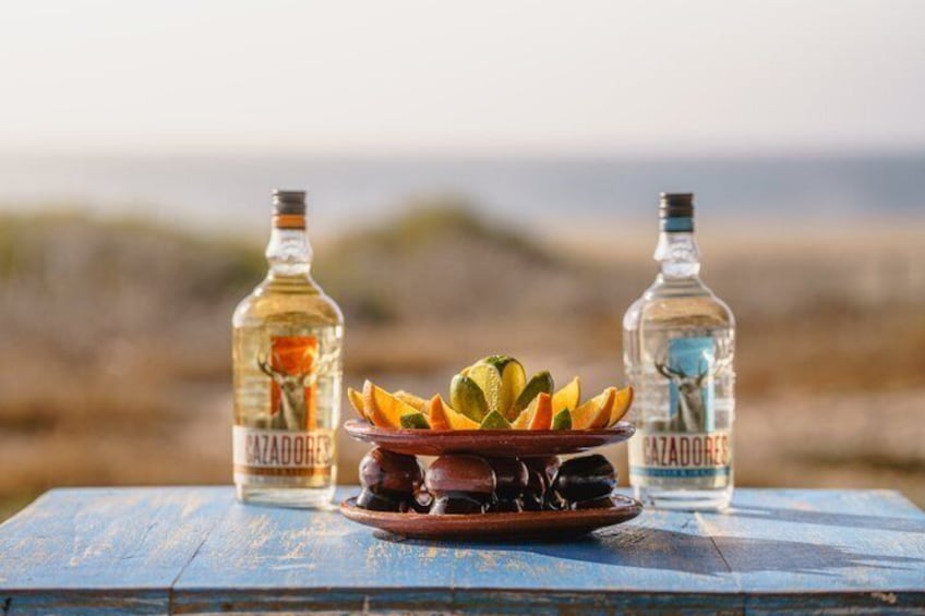 A table with two bottles of Cazadores tequila, a clay pot with fruit, and a coastal landscape in the background. 3