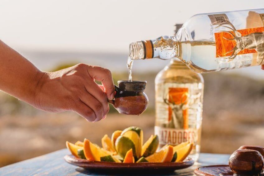 A hand pouring a clear liquid from a bottle into a small container, with a bottle of liquor and a plate of sliced fruit in the background. 4