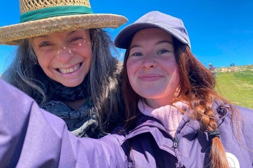 Two people wearing hats and jackets, with a clear blue sky and a grassy hill in the background. 2