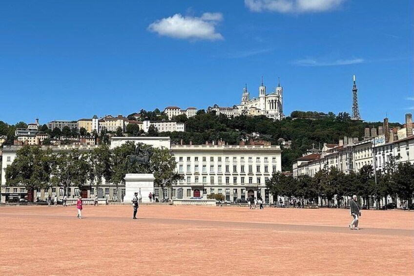 A wide open plaza with a large statue in the center, surrounded by historic buildings and a prominent church on a hill in the background. 2