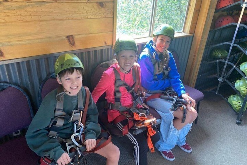 Three children in helmets and harnesses seated on a bench. 2