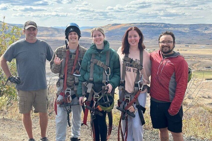 A group of people wearing harnesses and helmets, standing on a hill with a mountainous landscape in the background. 4