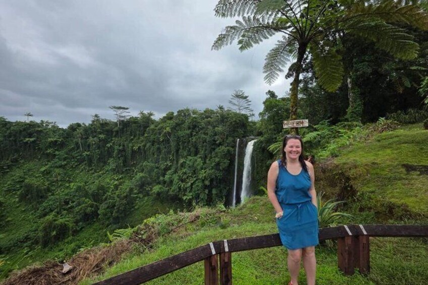 A person standing in front of a waterfall with lush greenery and a wooden fence. 4