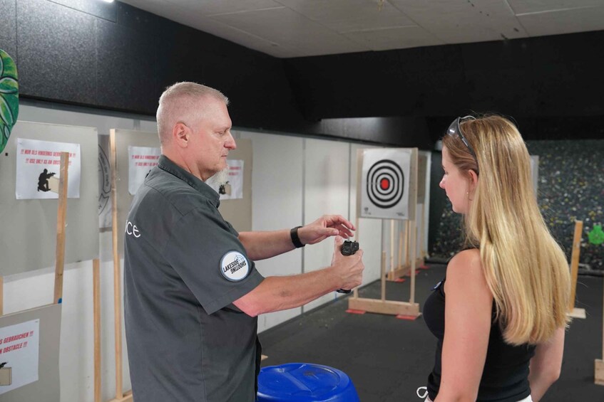 A man in a dark uniform with a badge, holding a small object, and a woman in a black top facing him. They are in an indoor setting with a target in the background. 1