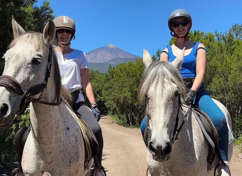 Two people horseback riding on a dirt path with a mountain in the background. 5