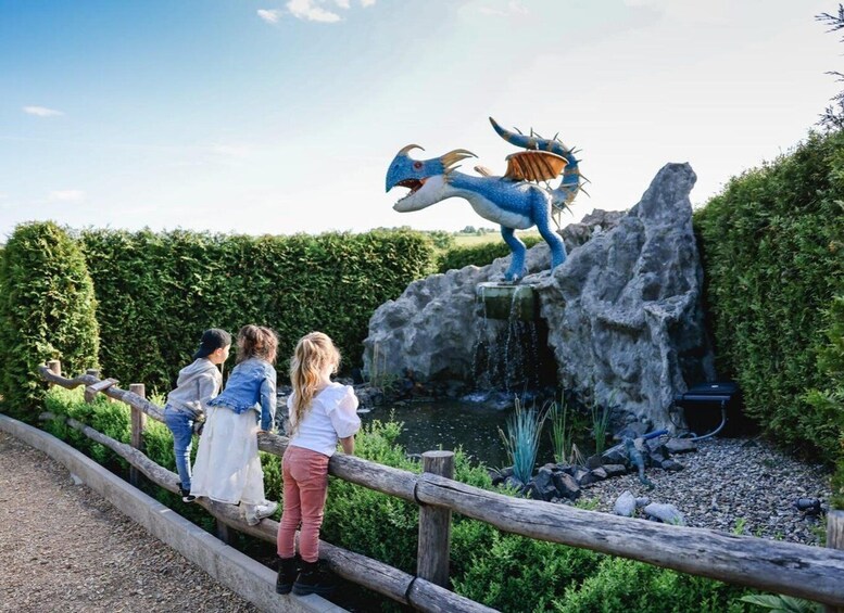 A group of children standing on a wooden fence, overlooking a rocky area with a water feature and a large blue dragon statue. 2