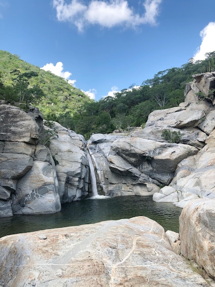 A waterfall cascading down a rocky cliff into a pool surrounded by lush green hills. 1