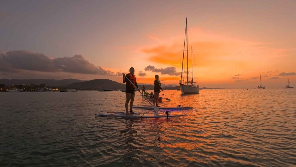 Two people paddleboarding on calm water with a sunset in the background and sailboats anchored nearby. 1