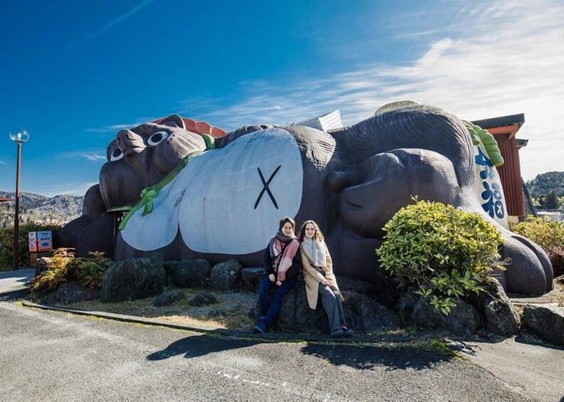 A large sculpture of a bear with a 'X' on its belly, two people sitting on a rock in front of it, and a clear blue sky. 2