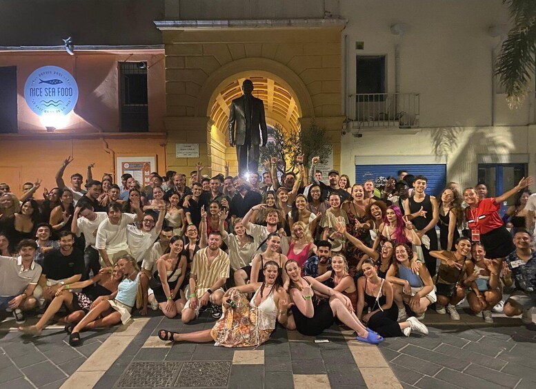 A large group of people posing for a photo in an outdoor setting with a building featuring an arched entrance and a sign that reads 'MICE SEA FOOD.' 1
