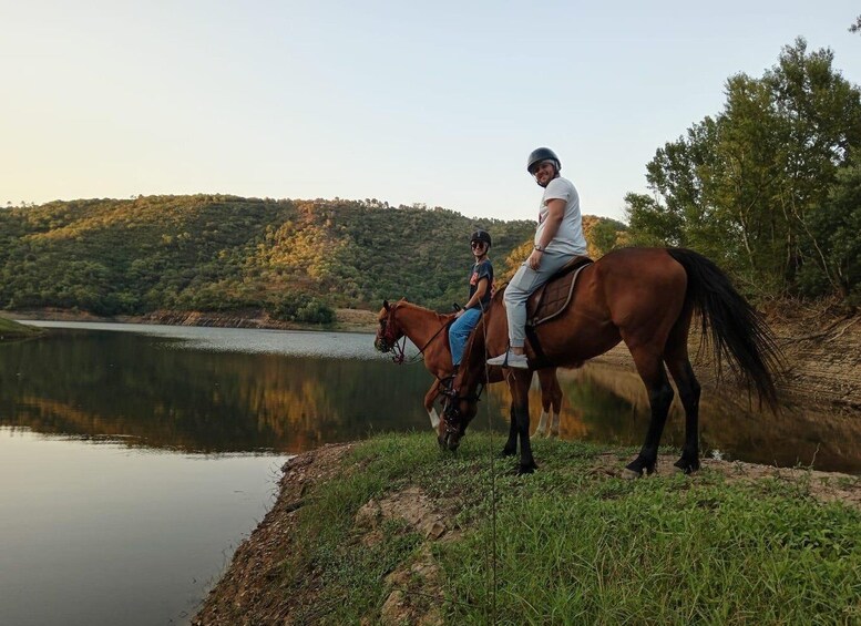 Two people riding a horse near a body of water with a forested hill in the background. 5