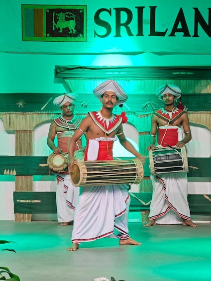 A cultural performance with musicians in traditional attire, a backdrop with the word 'SRILAN' and a lion emblem, and a stage with a green floor. 2