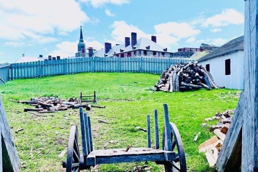 A grassy area with a pile of logs, a wooden fence, and a white building in the background. 4