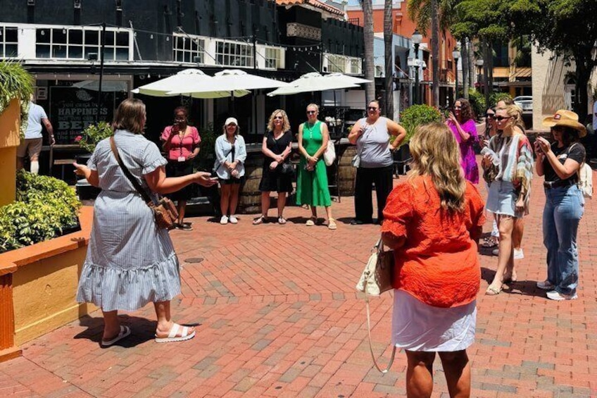 A group of people standing and conversing on a brick-paved outdoor area with umbrellas and a building in the background. 1