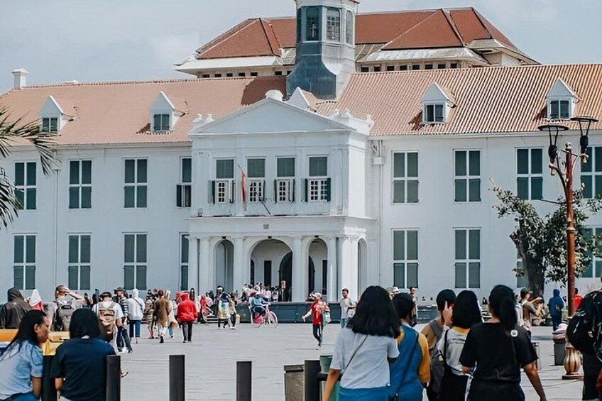 A large white building with a central entrance and multiple windows, surrounded by a crowd of people. 1