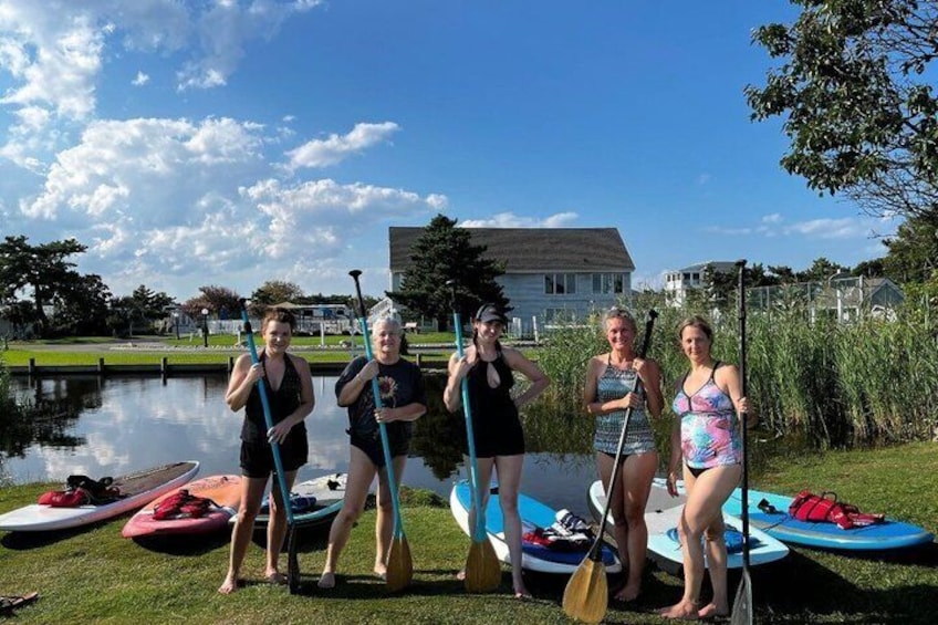 Four people standing by a body of water with paddleboards and paddles. 1