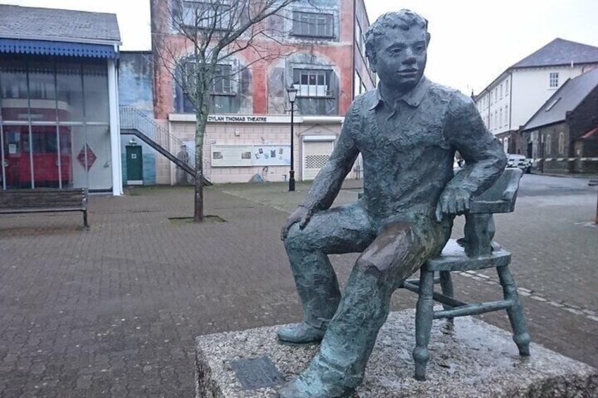 A bronze statue of a seated man on a bench in an urban setting with buildings and a tree in the background. 5