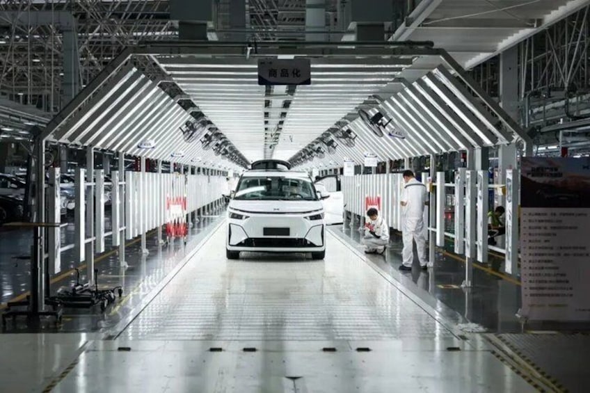 A car assembly line with a white vehicle in the center, workers on the right, and overhead lighting. 1